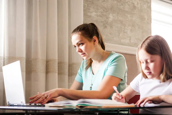 Young mom freelancer works from home remotely using a laptop, her daughter sits nearby and does homework. Horizontal photo