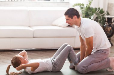 Genç baba ve küçük tatlı kızı evde ABS egzersizi yapıyorlar. Şirin bir çocuk ve babası kapalı alanda paspas eğitimi alıyor. Spor giyim bölümünde küçük bir bayan modelin odada kanepenin yanında egzersizleri var. Yatay fotoğraf