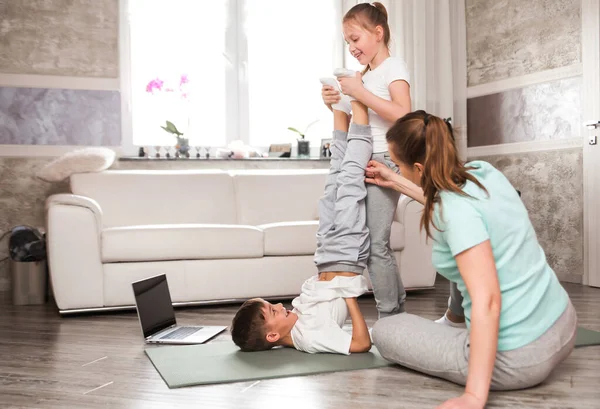 Mom daughter and son do gymnastic exercises together at home on the rug. Girl helps little brother to do birch stance. Horizontal photo