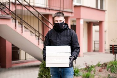 Siyah üniformalı, siyah koruyucu maskeli ve eldivenli yemek teslimatçısı eve beyaz kutularda yiyecek getiriyor. Yatay fotoğraf