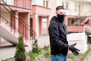 Siyah üniformalı, siyah koruyucu maskeli ve eldivenli yiyecek teslimatçısı eve çok katlı bir binanın dairesine beyaz kutularda yiyecek getiriyor. Yatay fotoğraf