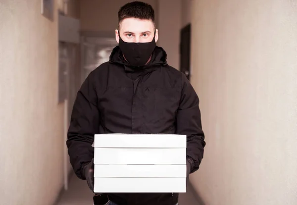 Food delivery man in black uniform, black protective mask and gloves delivers food to the house in boxes. Horizontal photo