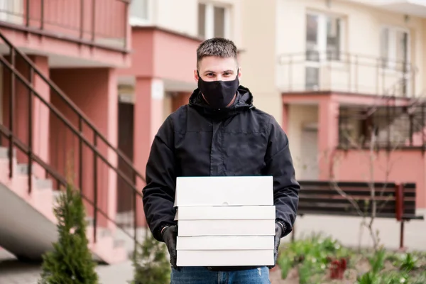 Food delivery man in black uniform, black protective mask and gloves delivers food to the house in white boxes. Horizontal photo