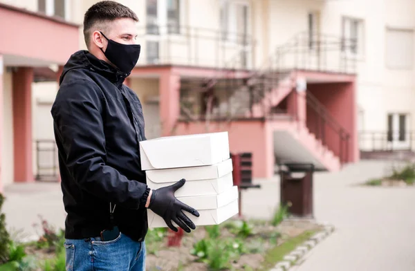 Food delivery man in black uniform, black protective mask and gloves delivers food to the house in white boxes.