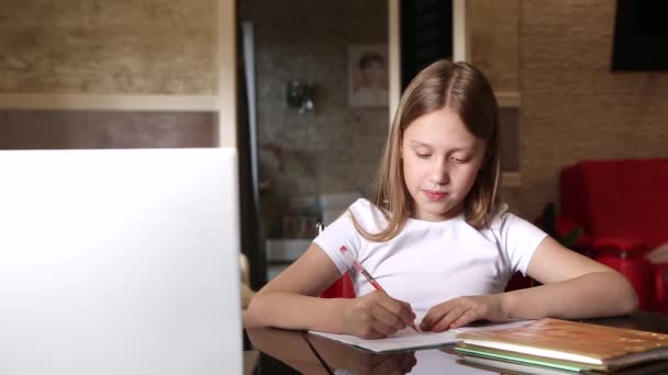 学校の年齢の少女は家で勉強している 女の子はノートパソコンのモニターでビデオレッスンを見て ノートブックにタスクを書きます ビデオ — ストック動画