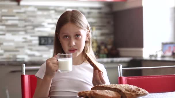 一个穿着白色T恤的女孩坐在厨房里 喝着杯子里的牛奶 吃着自家做的面包 — 图库视频影像
