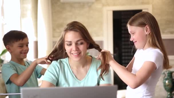一位年轻的女自由职业者在家里用笔记本电脑远程工作 女人的孩子帮她理发 — 图库视频影像