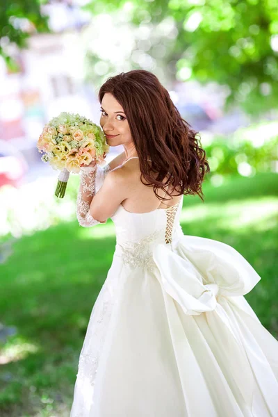 La hermosa novia sonriente con el pelo largo y fluido en magnífico vestido de novia blanco con un gran arco sosteniendo un ramo de flores en sus manos sobre un fondo soleado naturaleza verde . — Foto de Stock