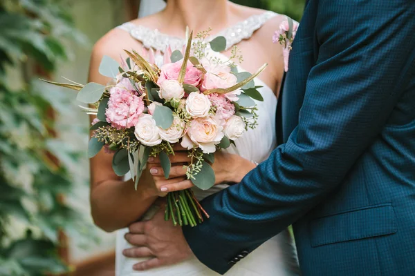 Bela noiva em um vestido branco segurando buquê de flores da primavera e seu noivo a abraça. Buquê de casamento. Momento de amor . — Fotografia de Stock