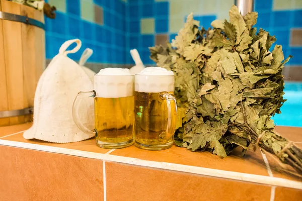 Bath accessories in the Russian bath. Bathroom items of traditional Russian sauna. two mugs of light beer, bathing caps, bath brooms from oak leaves against the background of the pool. — Stock Image Bath accessories in the Russian bath. Bathroom items of traditional Russian sauna. two mugs of light beer, bathing caps, bath brooms from oak leaves against the background of the pool. Royalty Free Stock Images