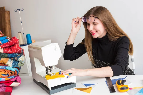 A bela menina sorridente com óculos na máquina de costura costurar pedaços de tecido colorido para colcha de retalhos, o processo de criação de cobertores scrappy . — Fotografia de Stock