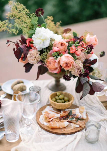 Wedding decor. Perfectly decorated holiday table for two with beautiful flower composition of fresh flowers in a vintage vase, red candles, thinly cut jamon, green olives and fresh bread. — Stock Photo, Image