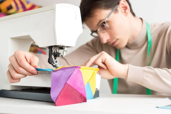 Jonge mannelijke op maat in de tabel werkt op de naaimachine en stitch ontwerper lappendeken van stukjes gekleurde doek. — Stockfoto