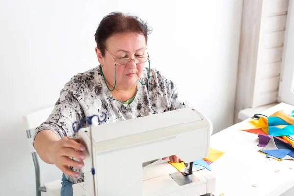 Retalhos e colchas na oficina de uma mulher alfaiate no fundo branco mulher alfaiate idoso com óculos costura na máquina de costura pedaços de tecidos coloridos . — Fotografia de Stock