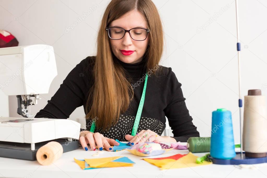 needlework and quilting in the workshop of a young woman, a tailor - pretty tailor in glasses at work with pieces of colored cloth on the table with threads, fabrics, needles and sewing machine.
