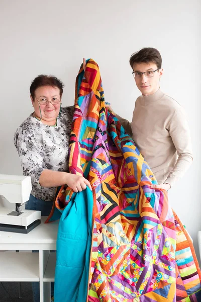 Naaien en quilten in het atelier van kleermakers - mannelijke en vrouwelijke lachende kleermakers samengehouden in handen een grote heldere quilt, genaaid van kladjes van gekleurde stof op de naaimachine. — Stockfoto