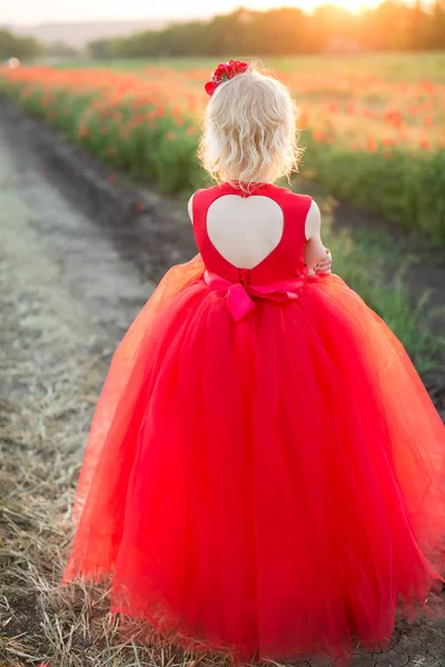 Девушка модель, детство, мода и летняя концепция - молодая блондинка в красном платье, стоящая спиной на дороге рядом с маковым полем, на ее спине большой вырез в форме сердца . — стоковое фото