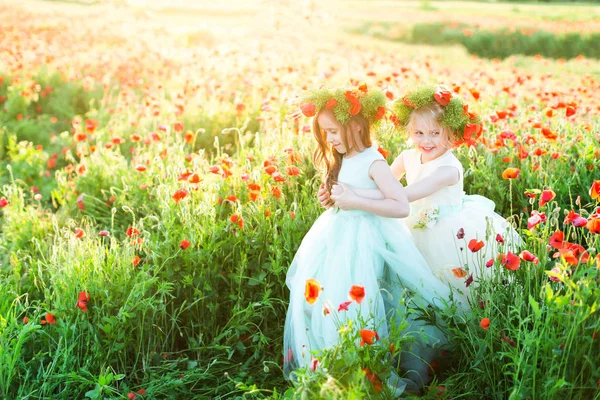 Modelo de niña, boda, amapolas, concepto de moda de verano - juego de dos niñas en vestidos de novia en un campo soleado de amapolas, las dos chicas riendo en coronas de flores de verano, cogidas de la mano . —  Fotos de Stock