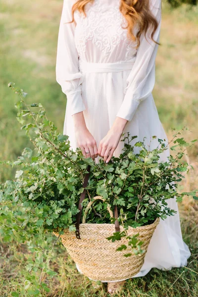 people, holidays and nature concept - young woman with red hair in white holiday dress is holding a large basket of green branches for decoration, girl florist on the summer field.
