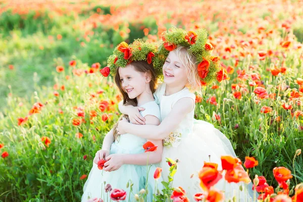 Modelo niña, boda, amapolas, concepto de moda de verano - dos niñas vestidas con vestido blanco y azul sonriendo en un campo de amapolas rojas, rubia abrazó a otro, en las cabezas de ellos coronas de amapola . — Foto de Stock