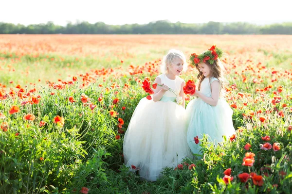 Маленькая девочка модель, детство, мода, летняя концепция - две маленькие подружки в белых и синих платьях, смеющиеся на весеннем поле, руки с букетами маков, на голове маковый венок . — стоковое фото