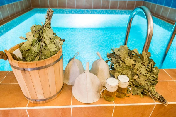 Badaccessoires im russischen Bad. Badartikel der traditionellen russischen Sauna. zwei Becher helles Bier, Badekappen, Besen aus Eichenblättern vor dem Hintergrund des Pools. — Stockfoto