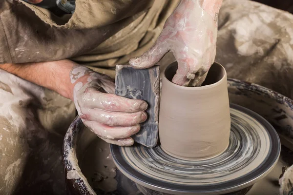 Создавая кружку из глины на крученом гончарном колесе. Грязные руки в глине и гончарные колеса с заготовкой. Скульптор в мастерской делает кувшин из глиняной посуды крупным планом . — стоковое фото