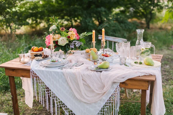 野餐、 夏天、 假日概念 — — 漂亮的装饰餐桌上绿色草坪与树木、 镂空白色桌布、 多彩花束牡丹的花和玫瑰，玻璃器皿，水果、 烛台、 柠檬水. — 图库照片