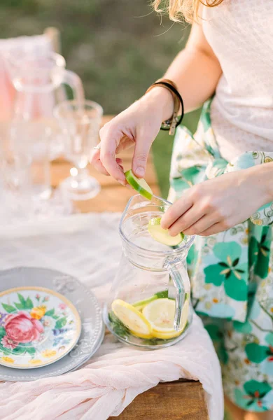 Picnic, gente, comida, verano, concepto de vacaciones: limonada de cóctel sin alcohol, manos de mujeres jóvenes mezclan rodajas de limón, lima y hojas de menta en un frasco de vidrio, mesa con platos festivos, vasos . — Foto de Stock