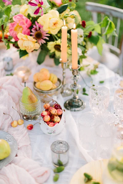 Pique-nique, nourriture, été, concept de vacances - gros plan sur une partie de la table décorée avec un beau bouquet de roses et pivoines roses, bougies allumées dans des chandeliers, fruits, verrerie, décor de glands . — Photo