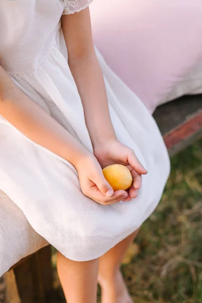 Personas, comida, concepto de vacaciones - dos manos de una niña pequeña en un vestido elegante blanco sosteniendo un albaricoque amarillo jugoso maduro, niña sentada en el banco con fondo de hierba de verano, vertical . — Foto de Stock