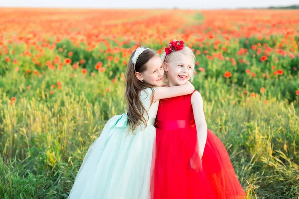 Девушка модель, детство, мода, природа и летняя концепция - две маленькие улыбающиеся девочки на поле из красного мака, они одеты в голубую и красную праздничную одежду, одна из них обняла вторую шею . — стоковое фото