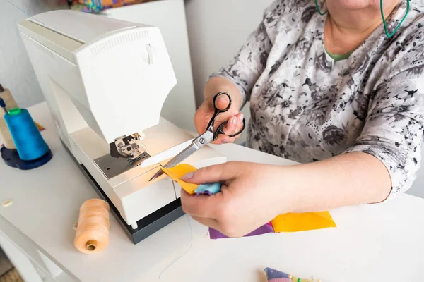 Bordado e colcha de mão na oficina de uma mulher alfaiate - close-up em mãos costureira trabalho na máquina de costura com tesoura, ela corta o fio sobre os tecidos coloridos costurados . — Fotografia de Stock