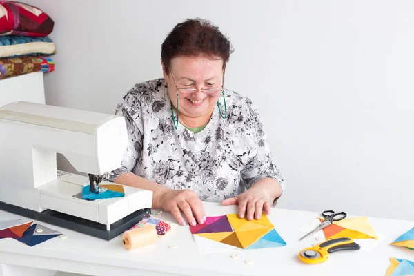 针线活和车间的裁缝女人-绗缝老人微笑妇女裁缝放在桌面上的有色的织物、 lscissors、 按钮、 别针、 刀具和缝纫机螺纹件 — 图库照片