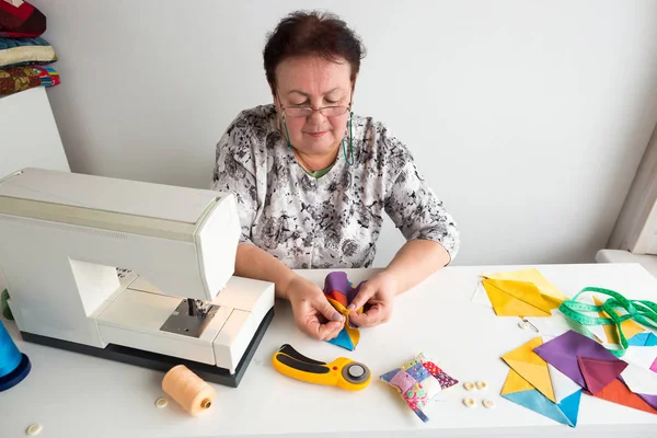 Costura y acolchado: el sastre más viejo cose manualmente piezas de telas brillantes en la mesa con una máquina de coser, hilo, agujas, alfileres, cojín, cinta métrica y botones . — Foto de Stock