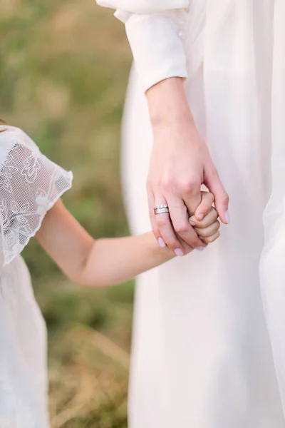 Novia, la infancia, el concepto de fiesta de boda - la mano de una niña sosteniendo la mano de una mujer joven adulta con anillos de boda en los dedos, la niña y la novia vestida con vestidos de fiesta blancos . — Foto de Stock