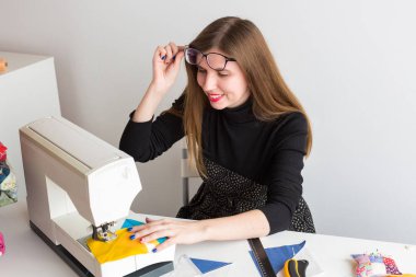 genç gülümseyen terzi yüzünde gözlük ayarlar, iş parçacığı ve yastık, döner bıçakları skeins tablo sonraki satırdaki dikiş patchwork bir dikiş makinesi ile beyaz masa başında oturur.