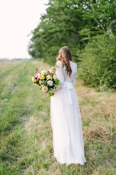 Ramo, gente, vacaciones de flores, regalo y concepto de arreglo floral hermosa hembra en un vestido blanco de moda posando con su espalda con ramo de flores frescas en el fondo de los árboles . — Foto de Stock