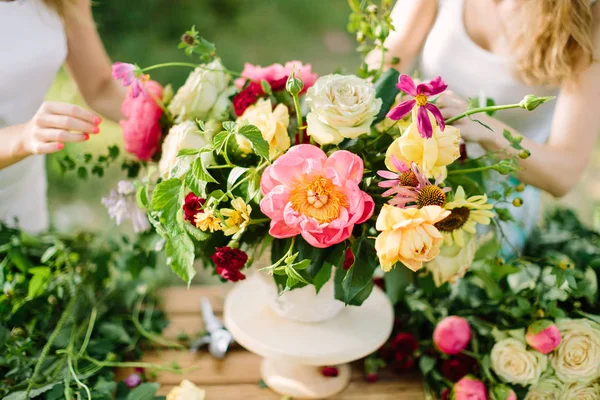 Boeket, mensen en bloemen regeling concept - handen van twee jonge vrouw opstelling van een boeket van mooie rozen roze pioenrozen, madeliefjes in witte vaas, kunst workshop bij houten tafel in de zomertuin. — Stockfoto
