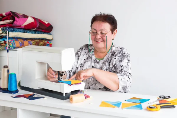 Retalhos e colchas na oficina de uma mulher alfaiate no fundo branco mulher idosa com óculos costura na máquina de costura restos de tecido colorido para retalhos . — Fotografia de Stock