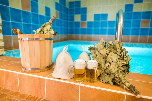 Bath accessories in the Russian bath. Bathroom items of traditional Russian sauna. two mugs of light beer, bathing caps, bath brooms from oak leaves against the background of the pool.