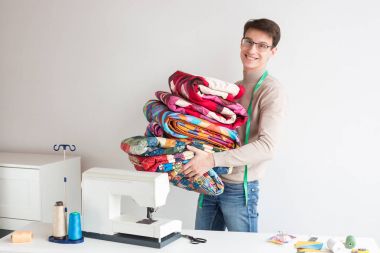 patchwork ve kapitone - dikişli renkli kumaş yığınını gülümseyerek terzi erkek taşır, o tablo bir dikiş makinesi, ölçüm bandı asılı boyun ile yakın.