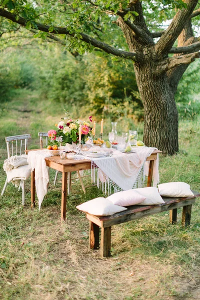 Piknik, léto, dovolená concept - sváteční stůl nastavení mezi velké stromy, prolamované ubrus, dřevěné lavice, židle, barevné kytice, džbán limonády, ovocné talíře, svícny, Selektivní ostření. — Stock fotografie