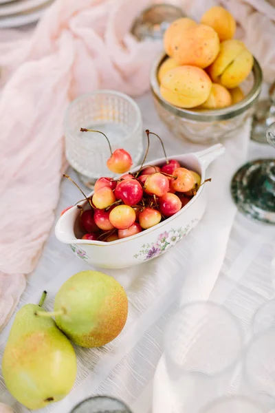 picnic, food, holiday concept - delicious fresh red and yellow cherries, pears, apricots on decorated table for the picnic, fruit plates and dishes, vegan food, white tablecloth, clean fresh fruit.