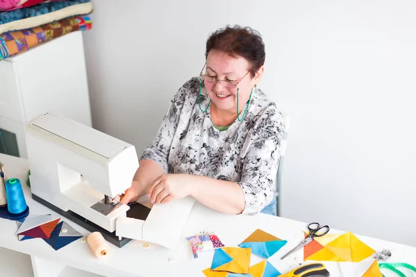 Patchwork en quilten in de werkplaats van een kleermaker vrouw op witte achtergrond - lachende vrouw met glazen voegt de draad in de naald van de naaimachine — Stockfoto