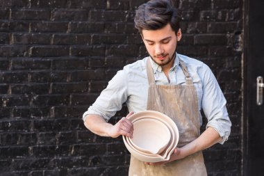 önlük içinde gülümseyen erkek ustası kil ürünleri, örneklerini gösterir yeni tutan usta Potter, mutfak eşyaları, Seramik Sanat kavramı - çanaklar, koyu gri tuğla duvar arka plan üzerinde mutlu ana bitmiş.