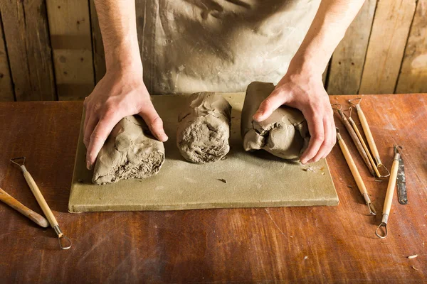 Cerâmica, argila, conceito de arte cerâmica - close-up em mãos de jovem mestre com os grandes pedaços de barro de fogo, macho vestido de avental, ceramista com matérias-primas em mesa de madeira com conjunto de ferramentas de escultura . — Fotografia de Stock