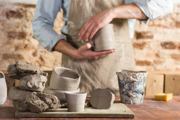 Cerâmica, oficina, conceito de arte cerâmica - mãos jovens artesãos segurando jarro não cozido, ceramista masculino stand no local de trabalho e vestindo avental, local de trabalho com fireclay e copos inacabados, foco seletivo . — Fotografia de Stock