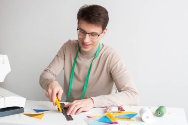 kapitone ve patchwork atölye - genç erkek oturma masada terzi ve renkli kumaş dikiş patchwork öğeler, bobinler iplik ve kumaş artıkları yanında yatıyordu için parçalarını kesip.