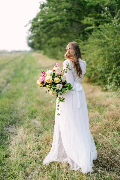 Casamento, casamento, beleza, natureza, design floral, conceito de país silhueta macia de noiva jovem em vestido branco nevado com cabelo castanho fluindo e grande buquê de peônias e rosas macias — Fotografia de Stock